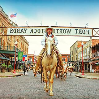 一名身着传统西部服装的牛仔骑着一匹棕白相间的马，站在“沃斯堡畜牧场”的标志下，标志横跨一条两旁是西部老建筑的街道